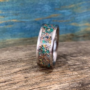 Pu&ograve; includere: Un anello d'argento con una fascia larga che presenta un'intarsio colorato di particelle macchiate di verde, oro e marrone.