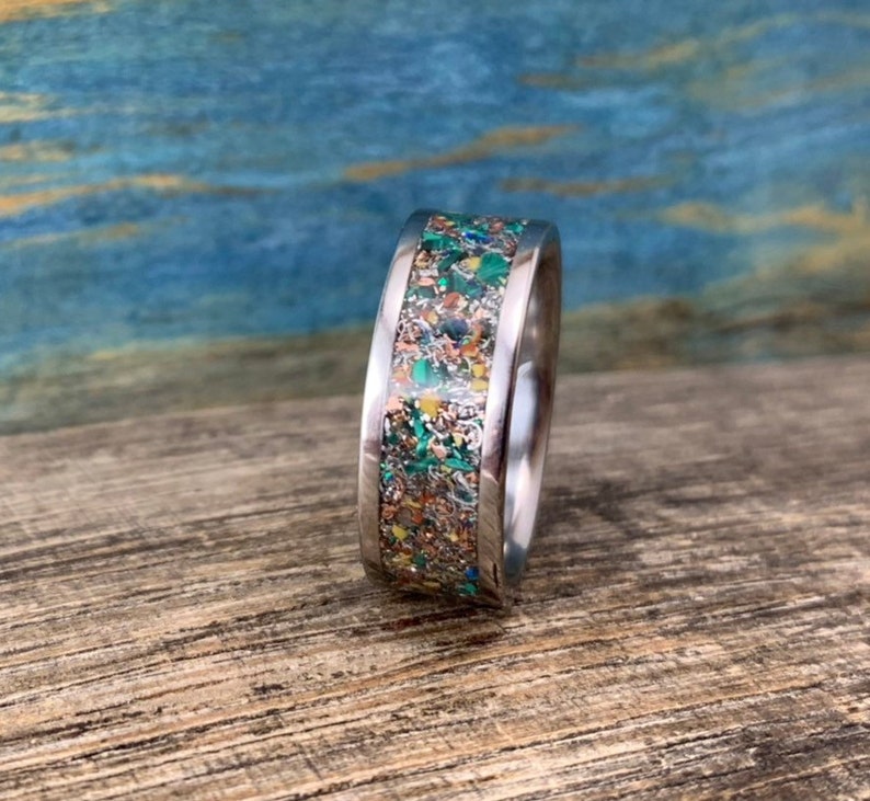Pu&ograve; includere: Un anello d'argento con una fascia larga che presenta un'intarsio colorato di particelle macchiate di verde, oro e marrone.