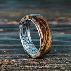 A zebrawood ring with a silver interior featuring an engraved floral pattern is angled on a dark wooden surface.