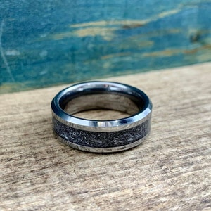 May include: A silver tungsten carbide ring with a beveled edge and a black inlay. The inlay has a textured, metallic finish.