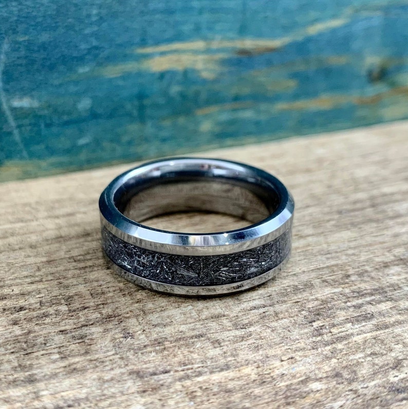 May include: A silver tungsten carbide ring with a beveled edge and a black inlay. The inlay has a textured, metallic finish.