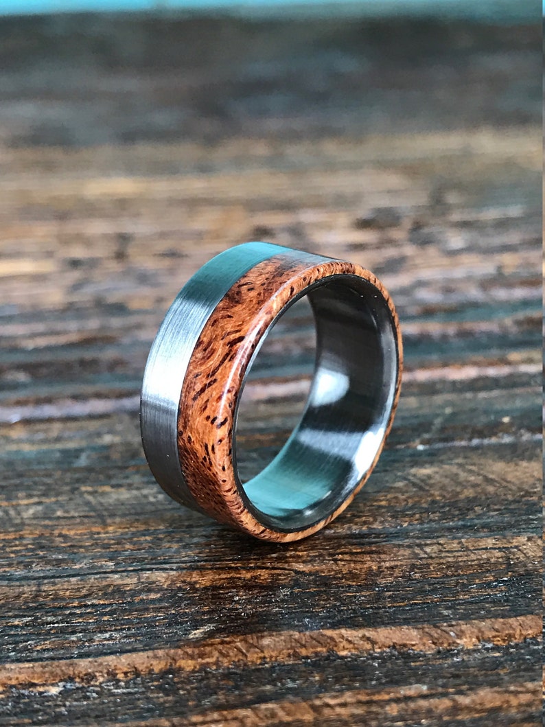 May include: A silver and wood ring with a brown wood inlay. The ring is on a wooden surface.
