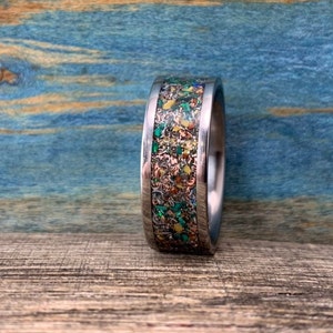 Pu&ograve; includere: Un anello d'argento con un'intarsio colorato di macchie verdi, gialle e arancioni. L'anello &egrave; su una superficie di legno.