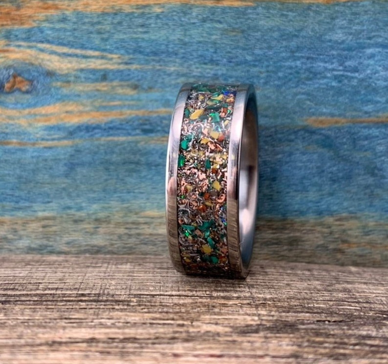 Pu&ograve; includere: Un anello d'argento con un'intarsio colorato di macchie verdi, gialle e arancioni. L'anello &egrave; su una superficie di legno.