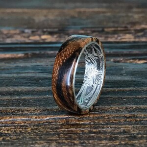 A zebrawood ring with a silver interior and engraved floral pattern is shown from a slightly different angle on a dark wooden surface, highlighting the wood's stripes.