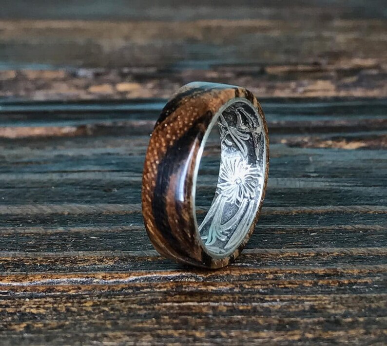 A zebrawood ring with a silver interior and engraved floral pattern is shown from a slightly different angle on a dark wooden surface, highlighting the wood's stripes.