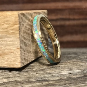 A gold ring featuring a colorful opal inlay rests vertically against a wooden block.