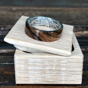 A zebrawood ring with a silver interior and engraved floral pattern rests on a light-colored wooden box with the lid slightly ajar.