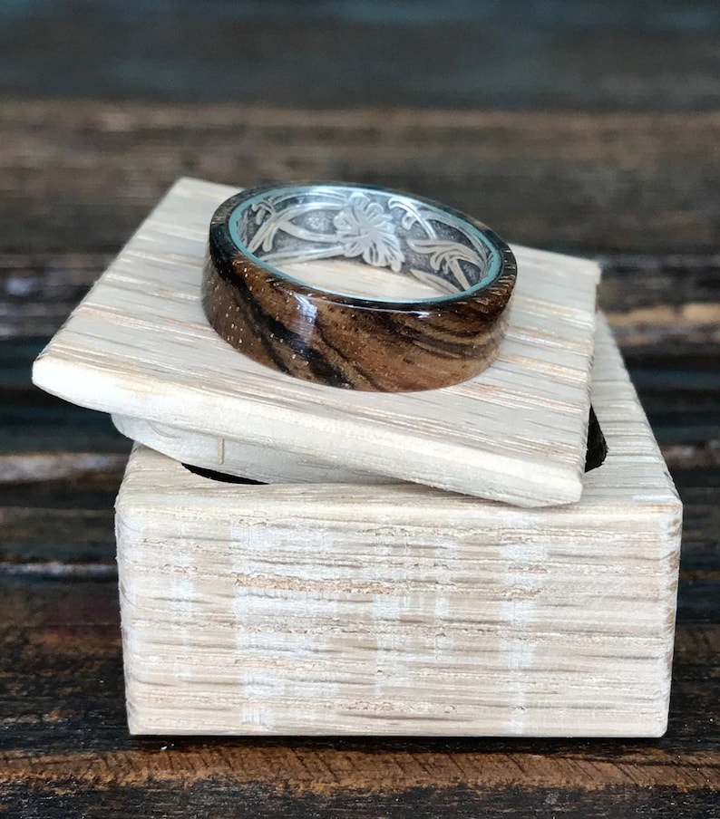 A zebrawood ring with a silver interior and engraved floral pattern rests on a light-colored wooden box with the lid slightly ajar.