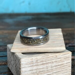 A silver ring with a gold fleck inlay sitting on top of a small open wooden box filled with white paper shred.