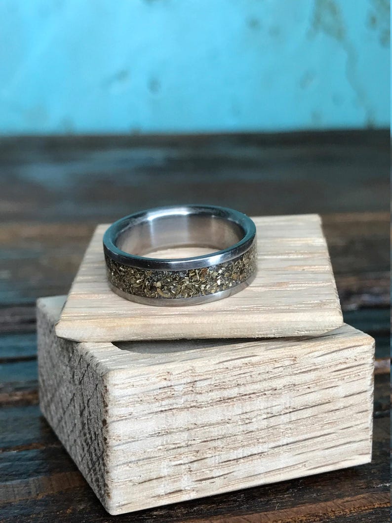 A silver ring with a gold fleck inlay sitting on top of a small open wooden box filled with white paper shred.