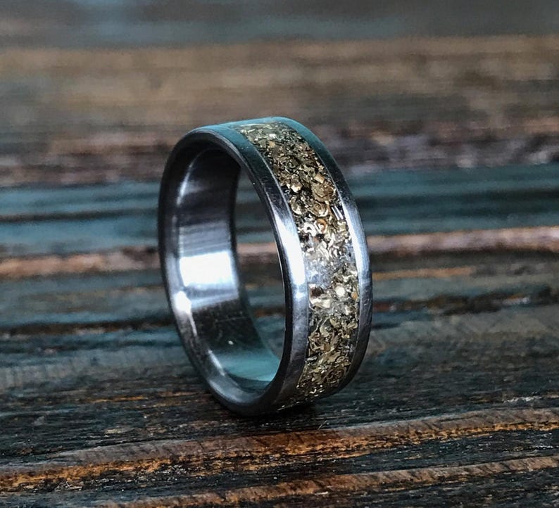 A silver-colored ring featuring a central inlay of metallic gold flakes, angled on a dark wooden surface.