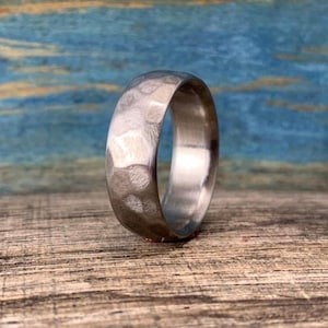 May include: A silver-colored ring with a hammered texture. The ring is set against a wooden surface with a blue and green background. The ring's surface reflects light, highlighting its textured design.