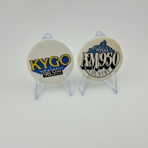 May include: Two vintage KYGO radio station pin badges. One badge features the KYGO logo in yellow and blue with "98.5 FM" text. The other badge has "KYGO AM950 COUNTRY" text in black and white.