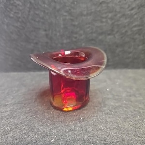 May include: A miniature top hat figurine crafted from translucent deep red glass. The object features a wide, curved brim and a cylindrical crown, with light reflecting off the smooth, polished surface of the glass material throughout the piece.