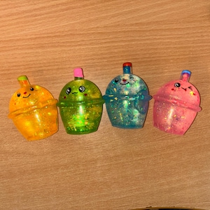 May include: Four glittery, colourful bubble tea-shaped containers with smiling faces. The containers are yellow, green, blue, and pink, each with a different coloured lid. The containers are on a wooden surface.