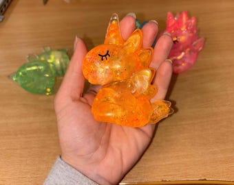 May include: A small, orange unicorn figurine with glitter embedded in the resin. The unicorn has closed eyes and a small horn.