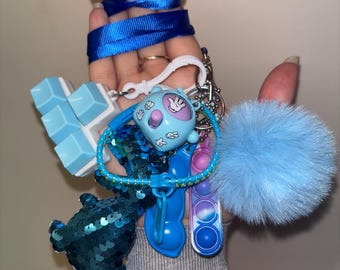 Puede incluir: Una colección de accesorios azules, que incluyen una figura con lentejuelas, un pompón esponjoso, un pequeño juguete pop-it y un llavero de personajes de dibujos animados. Una cinta azul está envuelta en la parte superior. Los artículos están unidos a un llavero plateado.