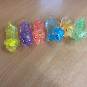 May include: A selection of colourful, glitter-filled dachshund figurines. The dachshunds are in various colours, including yellow, purple, red, blue, orange, and green. Each figurine has a small, round head and a long body. They are arranged on a light brown wooden surface.