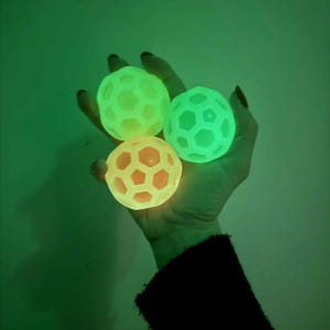 May include: Three glowing, spherical stress balls with a honeycomb design are held in a hand. The balls emit a bright, neon green and orange light. The background is a solid, dark green.