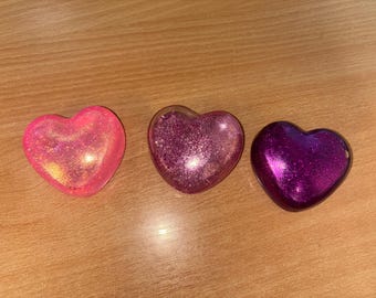 Puede incluir: Tres objetos en forma de corazón con purpurina. Los corazones son en tonos rosa y morado. El corazón de la izquierda es rosa brillante con purpurina dorada. El corazón del medio es rosa más oscuro con purpurina plateada. El corazón de la derecha es morado oscuro con purpurina plateada.