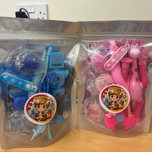 May include: Two clear plastic bags filled with colourful toys. The bag on the left contains a blue robot, a blue and white fidget toy, and a blue and white spinner. The bag on the right contains a pink giraffe, a pink and white fidget toy, a pink and white spinner, and a pink and white hair brush. Both bags have a sticker with a cartoon boy and the words "Happy Toys".