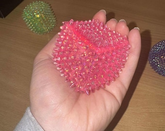Puede incluir: Una bola antiestrés rosa en forma de corazón con protuberancias puntiagudas. La bola se sostiene en una mano, con otras bolas antiestrés de diferentes colores visibles en el fondo. La bola antiestrés está hecha de un material translúcido.