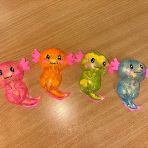 May include: Four colourful axolotl figurines with pink, orange, green, and blue glitter accents. Each figurine has a cute cartoon face with black eyes and a pink mouth.