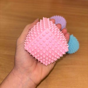 May include: A pink spiky ball toy with a textured surface. The ball is held in a hand.