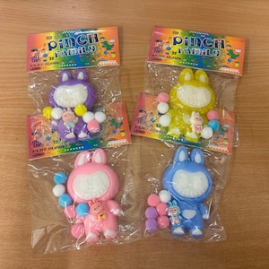May include: Four colourful bunny-shaped toys in plastic packaging. Each toy has a large white face, with a smaller bunny figure attached. The toys are purple, yellow, pink, and blue, with colourful ball-shaped accessories. The packaging reads "PINCH FAMILY".