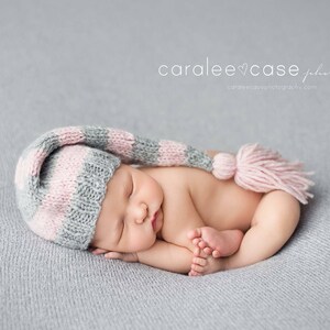 May include: A newborn baby wearing a striped pink, grey, and white knitted cap with a pink tassel, sleeping peacefully on a grey blanket. The baby is curled up, with tiny feet visible. The image has soft lighting.