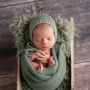 Op de afbeelding: Een pasgeboren baby is gewikkeld in een groene deken en draagt ​​een groene gebreide muts. De baby ligt in een witte houten doos met een groene, pluche voering.