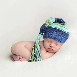 Peut inclure: Un nouveau-né portant un bonnet tricoté rayé bleu, vert et blanc avec un long pompon. Le bébé dort sur un fond blanc.