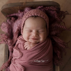 May include: A newborn baby wearing a pink knitted hat and swaddled in a pink blanket, smiling in a wooden crib.