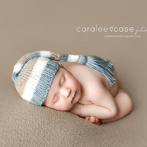 May include: A newborn baby wearing a striped blue, white, and tan knit cap with a tassel, curled up on a neutral-colored surface. The baby is sleeping peacefully. The image is a professional newborn photography shot.