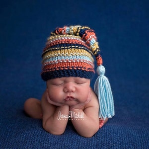 Puede incluir: Un bebé recién nacido con un gorro de punto a rayas azules, naranjas y amarillas con una larga borla azul. El bebé está acostado sobre una manta azul y tiene los ojos cerrados.