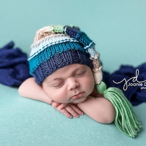 Op de afbeelding: Een pasgeboren baby draagt ​​een blauwe, groene en witte gebreide muts met een groene kwast. De baby slaapt op een blauwe en groene deken.