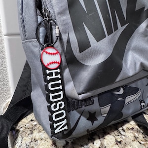 May include: Gray Nike backpack with a black swoosh logo and a black and white baseball-themed design. A white keychain with the name "HUDSON" and a red and white baseball is attached to the zipper.