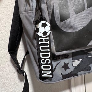 May include: Gray backpack with a black and white soccer ball keychain that says "HUDSON" in white letters.