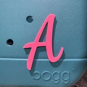 May include: A teal blue  Bogg bag with a pink letter A on the front. The bag has a textured surface and a logo that says "bogg" in a raised font.