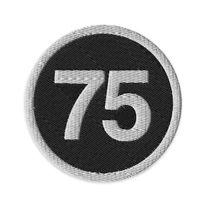 May include: Embroidered patch featuring the number 75 in white on a black circular background. The patch has a white border with a textured appearance. The design is simple and bold, suitable for various applications.