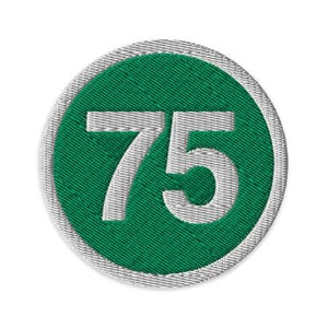 May include: A circular embroidered patch featuring the number 75 in white, set against a green background. The green circle is framed by a silver-grey border. The patch has a textured, stitched appearance.
