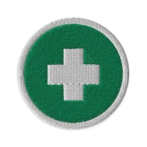 May include: Embroidered patch with a white cross on a green circle, edged with silver. The patch has a textured fabric appearance. The design is a well-known symbol, commonly linked to first aid or medical services, and is circular in shape.