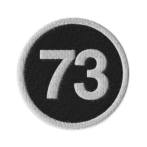 May include: Embroidered patch featuring the number 73 in white against a black circular background. The patch has a white border and is likely designed for application on clothing or accessories.
