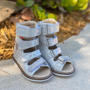 May include: A pair of silver orthopedic sandals with multiple adjustable straps. The sandals have a textured, glittery finish and are decorated with printed text. The soles are light brown, and the sandals are designed for children.