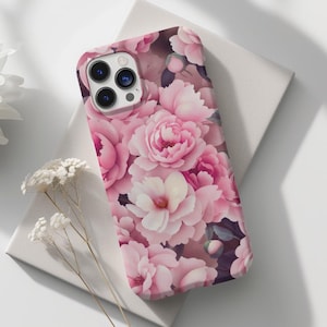 Puede incluir: Una funda de teléfono con un diseño floral rosa, con peonías y otras flores. La funda está sobre una superficie blanca, con una flor blanca y ramitas de flores secas. La funda del teléfono tiene un recorte para la lente de la cámara.