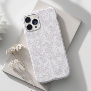 Puede incluir: Funda de teléfono con estampado floral blanco. La funda está sobre una superficie blanca con una flor blanca y ramitas de flores blancas. La funda del teléfono tiene un módulo de cámara con tres lentes.