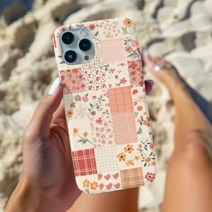 Op de afbeelding: Een telefoonhoesje met een bloemenpatchwork-ontwerp met verschillende patronen en kleuren, waaronder bloemen, harten en geruite vierkanten. De hoes heeft een crèmekleurige achtergrond en wordt in een hand gehouden.