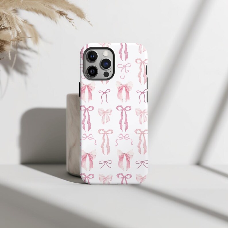 May include: A white phone case with a pink watercolor bow pattern. The case has a cutout for the camera lens.