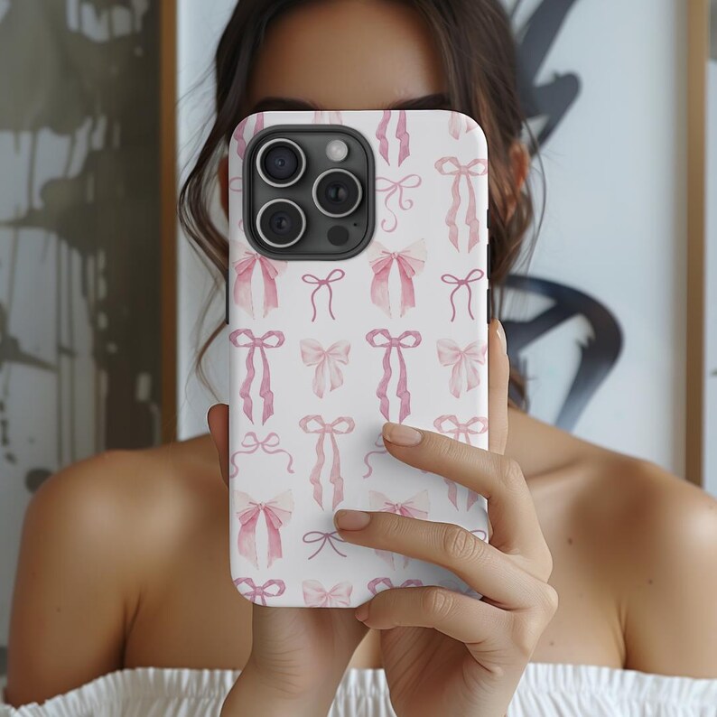 May include: A white phone case with a pink watercolor bow pattern. The case has a camera lens cutout and a white background.